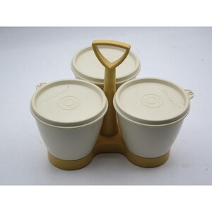 Puede incluir: Un servidor de condimentos vintage de Tupperware con tres recipientes de plástico color hueso y tapas a juego, unidos por un asa y una base central de plástico color amarillo mostaza. Cada tapa tiene grabado el nombre de la marca Tupperware.