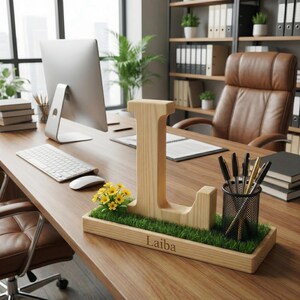 May include: A wooden desk organiser featuring a large letter L, a patch of artificial green grass, a cluster of small yellow flowers, and a black mesh pen holder. The wooden base is engraved with the name Laiba and holds several pens.