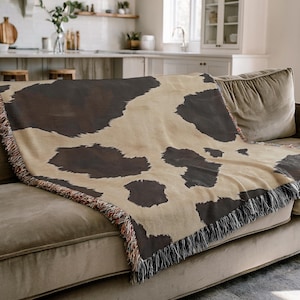 May include: A beige and brown cow print throw blanket draped over a brown sofa. The blanket has a fringe border with a mix of colors. The throw is made of a soft material and is a decorative home accessory.
