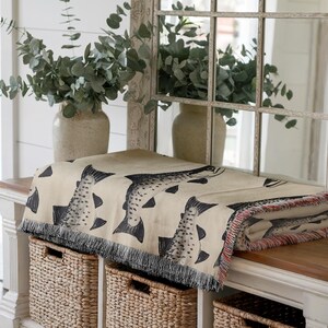 May include: A beige throw blanket with a repeating pattern of black fish, fringed edges, and red accents. The blanket is draped over a white bench with three woven storage baskets. A vase of greenery and a mirror are in the background.