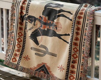 Op de afbeelding: Een beige geweven sprei met een zwart-bruine cowboy op een paard, een cactus en een geometrisch randontwerp. De sprei heeft een gefranjerde rand en is over een witte balustrade gedrapeerd.