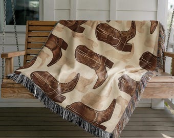 Op de afbeelding: Een beige sprei met een herhalend patroon van bruine cowboylaarzen. De laarzen zijn gedetailleerd met stiksels en schaduwen, tegen een lichtere, gevlekte achtergrond. De sprei heeft een gefranjerde rand.