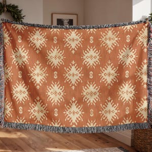 May include: A woven throw blanket with a repeating geometric pattern in cream and rust-orange. The blanket has a fringed edge in black and cream. The pattern features diamond and star-like shapes, creating a textured appearance. This blanket is suitable for home decor.