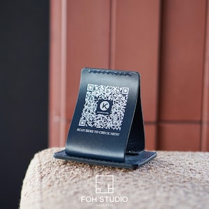 Pode incluir: Um suporte de mesa em couro azul marinho com um código QR e o texto "SCAN HERE TO CHECK MENU". O suporte é exibido em uma superfície bege. O logotipo "FOH STUDIO" está na parte inferior.