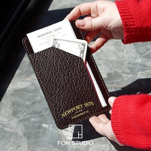 May include: A dark brown leather wallet with a textured finish, holding a business card and a five-dollar bill. The wallet has gold lettering that reads "NEWPORT 1875" and the FOH STUDIO logo.