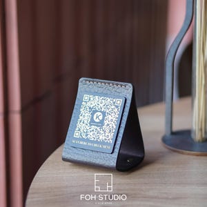 May include: A dark brown leather table stand with a QR code for a menu. The stand is angled and has a stitched border. The text "SCAN HERE TO CHECK MENU" is visible. A metal vase with dried stems is in the background.