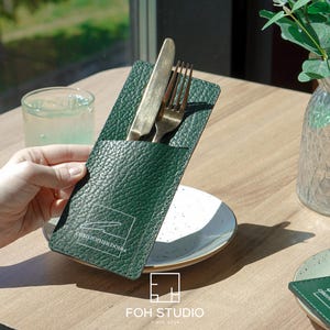 Könnte beinhalten: Dunkelgrüner Leder-Besteckhalter mit Messer und Gabel. Der Halter trägt die Aufschrift "CHRISTOPHER DOSSA" und "FOH STUDIO". Das Besteck ist goldfarben.
