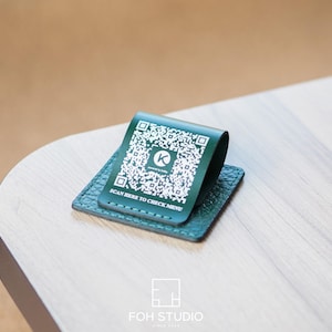 May include: A dark green leather QR code holder with a matching base, resting on a light wood surface. The holder displays a QR code and the text "SCAN HERE TO CHECK MENU". The holder is branded with a white "K" logo.