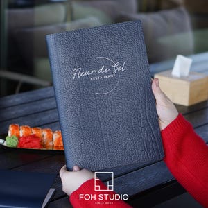May include: A dark blue, textured menu cover featuring the white printed text Fleur de Sel RESTAURANT with a circular graphic element. The rectangular cover has a pebbled surface finish and is held upright, displaying the front cover design clearly.