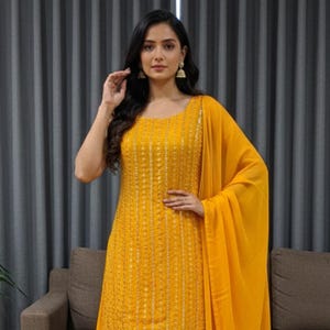 May include: A woman wearing a bright yellow Indian salwar kameez. The outfit includes a long tunic with vertical embellishments, matching wide-leg pants, and a flowing scarf. The text "Viorafashion studio" is in the bottom left corner.
