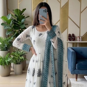 May include: A white floor-length dress featuring a round neckline, long sleeves with teal cuffs, and a repeating dark floral pattern. The garment includes a matching sheer teal-bordered dupatta draped over the shoulder with intricate detailing.