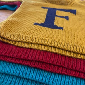 Peut inclure: Pull en tricot jaune moutarde avec un grand "F" bleu marine sur le devant. Le pull est empilé avec des tricots rouges et turquoise pliés. Le tricot est fait d'une matière douce et confortable.