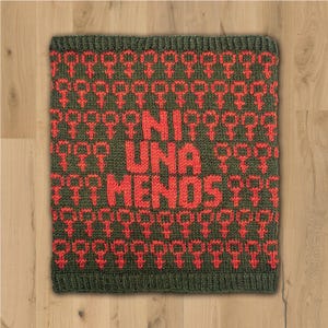 Op de afbeelding: Een gebreid vierkant in groen- en roodtinten. Het ontwerp bevat herhaalde rode symbolen en de woorden "UNA MENOS" in het rood. Het item wordt gepresenteerd op een lichtbruin houten oppervlak.