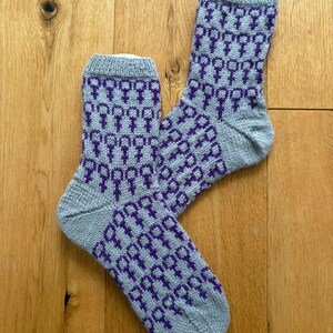 Könnte beinhalten: Ein Paar graue Strickstrümpfe mit einem sich wiederholenden lila Muster. Das Muster ist ein stilisiertes Symbol. Die Socken sind aus einem weichen, strukturierten Material gefertigt und haben einen gerippten Bund.