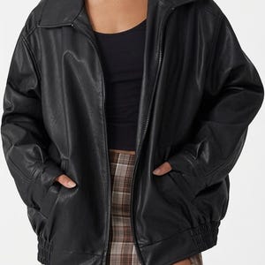 Pode incluir: Casaco preto de couro sintético com fecho de correr, gola e bolsos. O casaco tem um corte folgado com punhos e bainha elásticos. A modelo usa um top preto e uma saia xadrez.