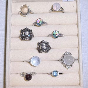 May include: A collection of silver rings with various gemstones displayed in a beige jewelry tray. The rings feature stones in shades of pink, blue, and iridescent colors. Some rings have ornate designs, including a cat face.