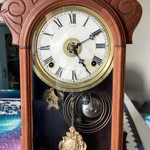 May include: A wooden mantel clock with a carved top, featuring a white circular face with black Roman numerals and hands. The lower section has a glass panel revealing internal brass gears, a coiled spring, and a decorative gold-toned pendulum.