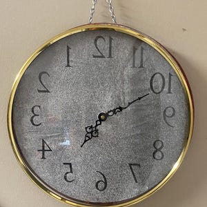 May include: A round wall clock with a gold-toned frame and a silver glitter-textured face. The clock features black Arabic numerals arranged in reverse order, black ornate hands, and is suspended by a silver metal chain attached to the top.