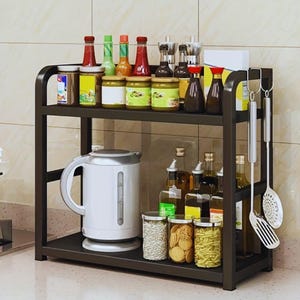 Puede incluir: Un estante de cocina de metal negro de dos niveles lleno de varios frascos, botellas y una tetera eléctrica blanca. El estante superior contiene condimentos, mientras que el estante inferior tiene frascos de comida y utensilios de cocina.