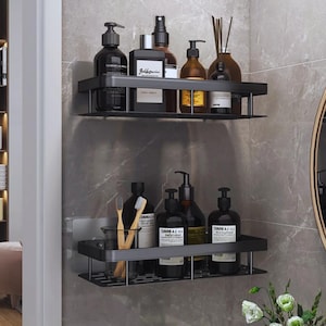 May include: Two black metal bathroom shelves fixed to a grey tiled wall. The shelves display various dark-coloured toiletries, including soap dispensers, spray bottles, and reed diffusers. A toothbrush and a glass are also visible.