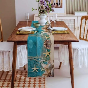 Op de afbeelding: Een houten eettafel met een strandthema tafelloper. De loper heeft een ontwerp van zeesterren, schelpen en een nautisch net tegen een blauw en beige achtergrond. De tafel is gedekt met borden, glazen en servetten.