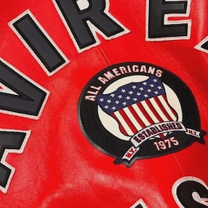 May include: Close-up of a red leather jacket with black and white lettering spelling out "FIRE" and a circular patch. The patch features an American flag design with the words "ALL AMERICANS ESTABLISHED 1975 N.Y."