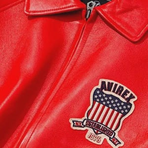 May include: A close-up of a red leather jacket with a collar and zipper. The jacket features a patch with the text "AVIREX" above a design of the American flag and the text "NY ESTABLISHED 1975 NY".
