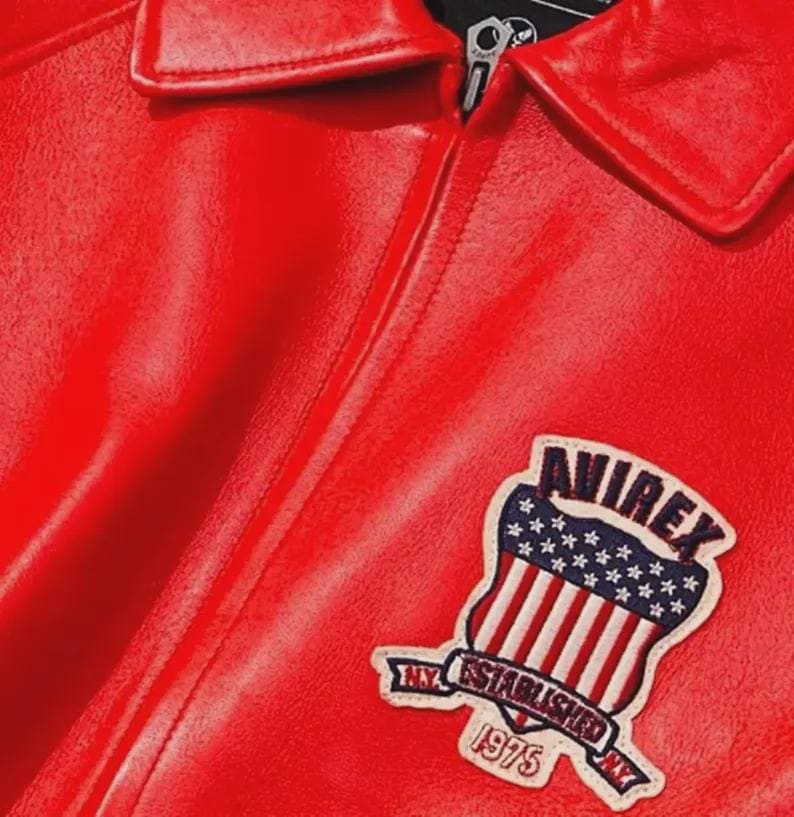 May include: A close-up of a red leather jacket with a collar and zipper. The jacket features a patch with the text "AVIREX" above a design of the American flag and the text "NY ESTABLISHED 1975 NY".