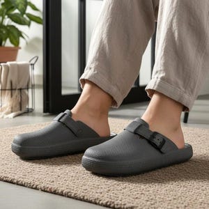 May include: Gray clogs with adjustable straps and buckle closures. The clogs have a textured surface and are worn indoors. The image shows the clogs on a beige rug.