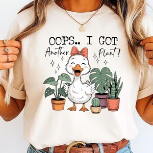 May include: Cream-colored t-shirt featuring a cartoon duck with a pink bow, surrounded by potted plants. The text "OOPS.. I GOT Another Plant!" is printed above the duck. The design includes stars and a smiling duck.