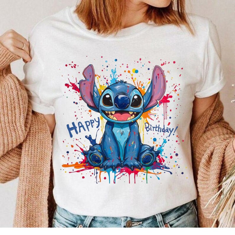 May include: White t-shirt featuring a blue cartoon character with large ears and the text "Happy Birthday!" in blue. The design includes colorful paint splatters. The shirt is a casual wear item.