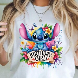 May include: White t-shirt featuring a blue cartoon character with large ears, adorned with flowers and butterflies. The design includes a banner that reads "BIRTHDAY GIRL!" with colorful paint splatter accents, and a cupcake.
