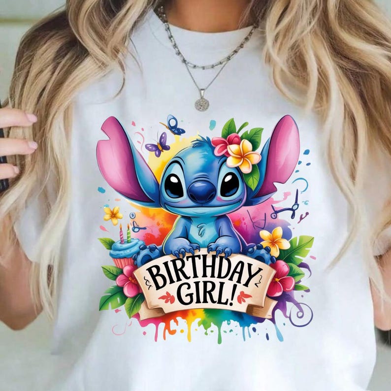 May include: White t-shirt featuring a blue cartoon character with large ears, adorned with flowers and butterflies. The design includes a banner that reads "BIRTHDAY GIRL!" with colorful paint splatter accents, and a cupcake.