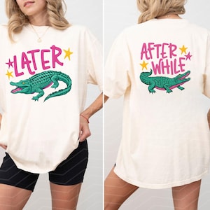 May include: Cream-colored t-shirt with a green alligator graphic and the word "LATER" in pink on the front. The back of the shirt features the words "AFTER WHILE" and a matching alligator graphic.