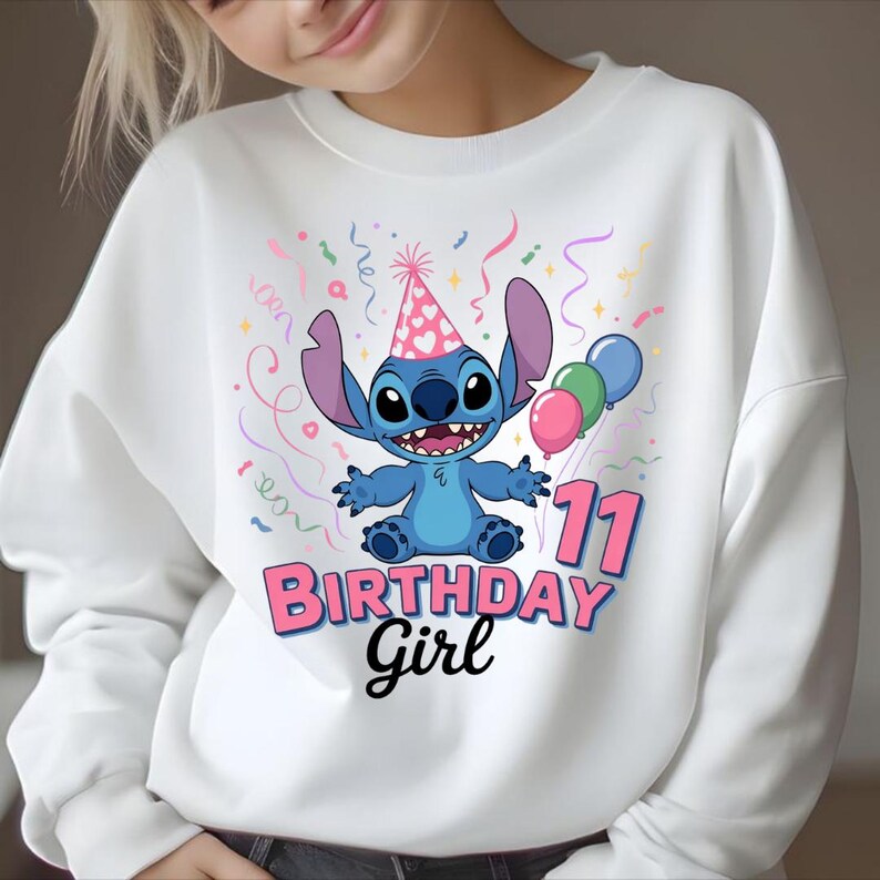 May include: White sweatshirt featuring a blue cartoon character wearing a party hat and holding balloons. The graphic includes the text "BIRTHDAY GIRL" and the number "11" in pink. Confetti and streamers surround the character.