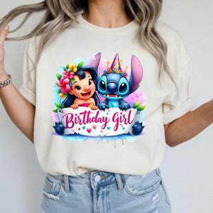 May include: A cream-colored t-shirt featuring a colorful graphic of two cartoon characters, one with a flower crown and the other with a party hat. A banner below reads "Birthday Girl" in pink script.