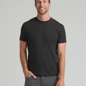 Op de afbeelding: Een zwart T-shirt met ronde hals. Het shirt met korte mouwen is gemaakt van een zacht materiaal. De persoon op de foto draagt een grijze broek. De achtergrond is effen lichtgrijs.