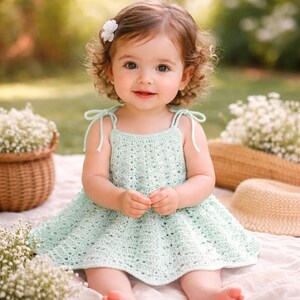May include: A light green crochet dress with tie straps. The dress is worn by a young child with curly brown hair and a white flower hair accessory. The child is sitting outside on a blanket.