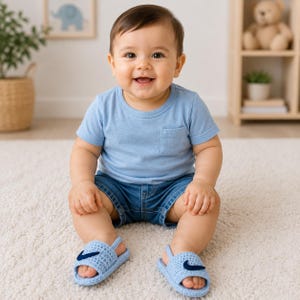 May include: A light blue short-sleeved t-shirt with a small chest pocket, dark blue denim shorts, and light blue crocheted sandals featuring a dark blue swoosh logo on the top strap. The clothing is worn by a smiling infant sitting on a white rug.