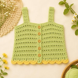 May include: A light green sleeveless crochet top featuring a vertical row of five wooden buttons down the center. The garment has a textured open-stitch pattern and a scalloped hemline finished in a contrasting pale yellow yarn color.