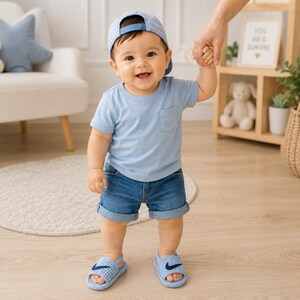 May include: A light blue t-shirt with a chest pocket, blue denim shorts with cuffed hems, and a light blue baseball cap. The outfit is completed with light blue rubber sandals featuring a dark blue logo on the top strap of each shoe.