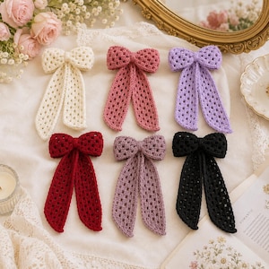 May include: Six crochet hair bows arranged in two rows. The top row features cream, dusty rose, and lavender bows. The bottom row includes deep red, mauve, and black bows. Each bow is crafted with a square-stitch pattern and has two long tails.