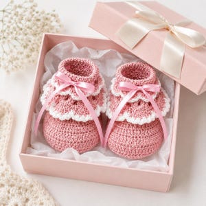 May include: Handmade pink crochet baby booties with white scalloped edging and pink ribbon bows, displayed in a pink gift box tied with a satin ribbon. The booties rest on white tissue paper.