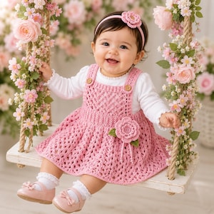 May include: A baby wearing a pink crocheted dress with a flower applique, a matching headband, and pink shoes. The baby is sitting on a swing decorated with pink flowers. The dress has wooden buttons and a white long-sleeved shirt underneath.