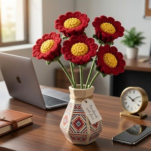 May include: A bouquet of five handmade crochet flowers in a decorative vase. The flowers have red petals and yellow centers. The vase is cream-colored with a geometric pattern in red and blue. A tag reads "Handmade with Love".