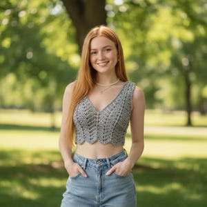 May include: A grey crochet crop top with a V-neck and a scalloped hem. The sleeveless top is likely handmade. The person is wearing blue jeans and a pearl necklace.