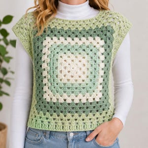 May include: A green and cream-colored crochet top with a square pattern. The sleeveless top is made of a soft yarn and has a round neckline. The top is worn over a white long-sleeved shirt and blue jeans.