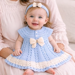 Può includere: Una bambina indossa un vestito all'uncinetto azzurro con un fiocco e rifiniture color crema. Il vestito ha una fascia abbinata con un fiocco. La bambina sta sorridendo.