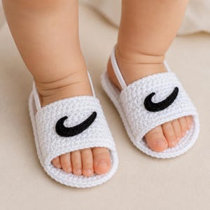 May include: A pair of white crocheted baby sandals featuring a black curved swoosh-like applique on the front strap. The sandals have a simple open-toe design with a thin white heel strap to secure them around the ankle, showcasing a textured yarn construction.