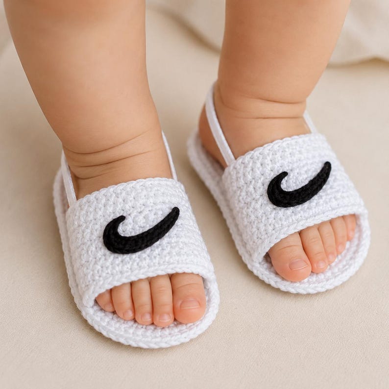 May include: A pair of white crocheted baby sandals featuring a black curved swoosh-like applique on the front strap. The sandals have a simple open-toe design with a thin white heel strap to secure them around the ankle, showcasing a textured yarn construction.